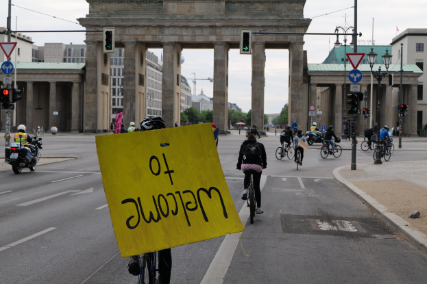 Eine Gruppe von Menschen mit Helmen fährt Fahrräder auf einer Straße vor dem Brandenburger Tor in Berlin, Deutschland, vorbei, während eine Person eine gelbe Tafel hält, Straßenlaternen, Verkehrszeichen, Gebäude, Bäume und einen klaren blauen Himmel im Hintergrund.
