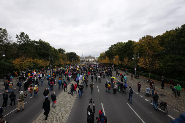 Große Gruppe von Menschen marschiert eine baumbestandene Straße mit Laternenpfählen entlang, hält Kameras, mit einem Gebäude und einem klaren Himmel im Hintergrund, was auf eine Demonstration in Berlin hinweist.