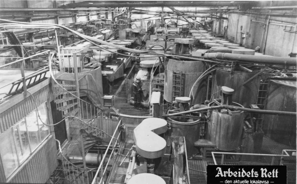 Schwarzes und weißes Foto einer großen Fabrikhalle mit einer Person zwischen Industriegeräten, Rohren und konstruktiven Elementen, mit Text am unteren Rand, der "Arbeidets Rett, die größte Fabrik der Welt" lautet.