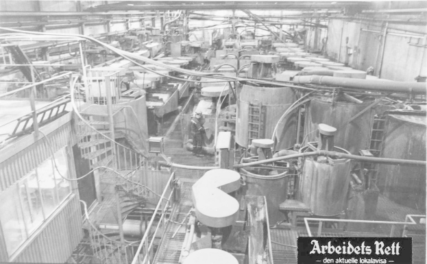 Schwarzes und weißes Foto einer großen Fabrikhalle mit einer Person zwischen Industriegeräten, Rohren und konstruktiven Elementen, mit Text am unteren Rand, der "Arbeidets Rett, die größte Fabrik der Welt" lautet.
