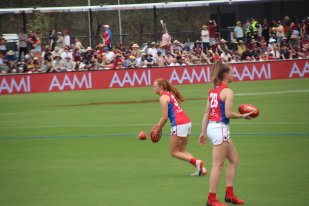 Zwei Frauen in Sportkleidung spielen Australian Football League auf einem Feld, eine hält einen Ball, mit Zuschauern und Werbetafeln im Hintergrund.