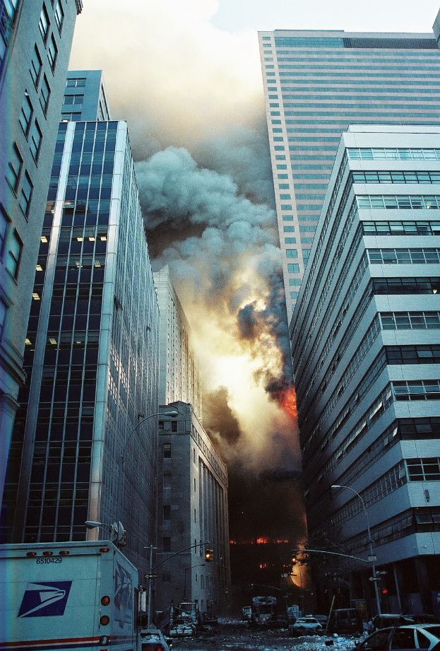 Eine belebte Stadtstraße mit Fahrzeugen, Laternenmasten und Gebäuden, mit Rauch und Feuer im Hintergrund.