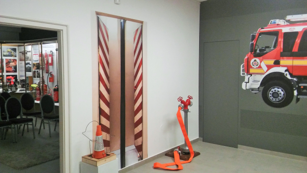 Ein Raum mit einer Wandmalerei eines Feuerwehrautos, einer Tür, einem Verkehrskegel, einem Ständer, einem Teppichboden, Stühlen, Tischen, Feuerlöschern und anderen Gegenständen.