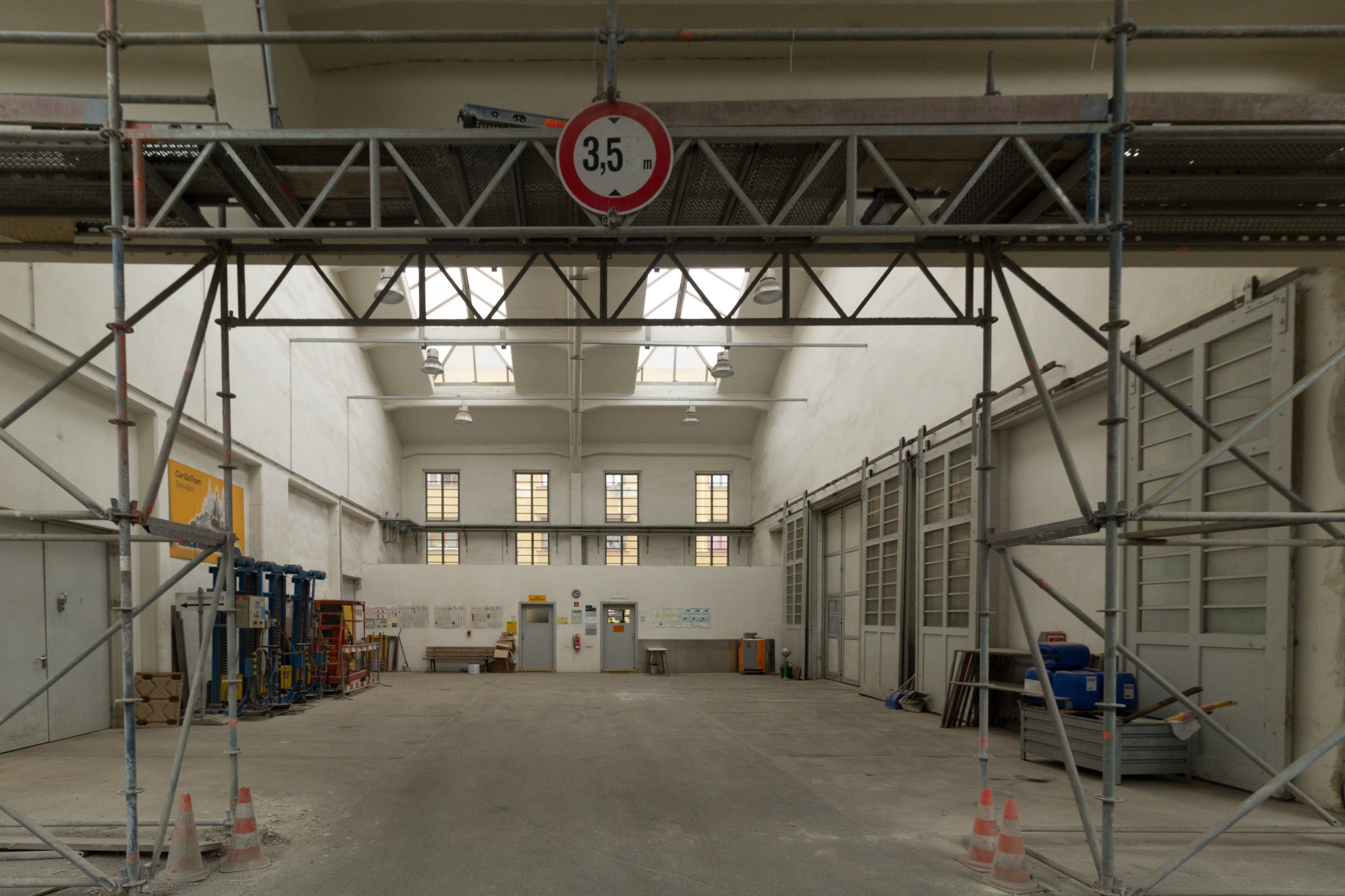 Lagerhalle mit Gerösten, Absperrgittern, Metallstangen, einem Schild, Maschinen, einer Tür, Fenstern, Wänden, Deckenleuchten und einem Dach.
