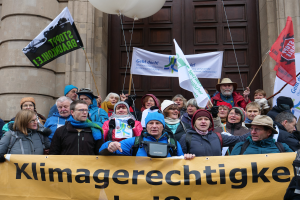 Eine Gruppe von Menschen steht vor einem Gebäude mit Säulen und einer Tür, hält ein Transparent mit Text, Fahnen und Ballons und nimmt an einem Klimastreik in Deutschland teil.