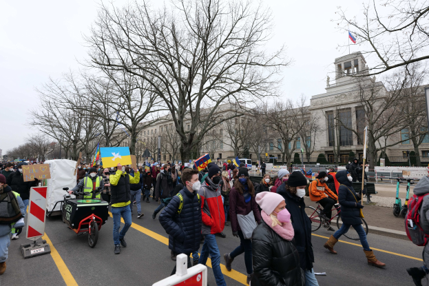 Eine große Gruppe von Menschen marschiert auf einer Straße bei einer Demonstration, einige halten Schilder und andere fahren Fahrräder, mit Bäumen und einem klaren blauen Himmel im Hintergrund.