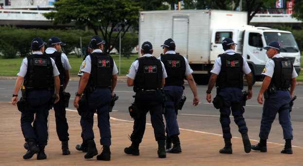 Eine Gruppe von Beamten in Uniform, einschließlich Mützen, Hemden, Schutzwesten, Uhren, Hosen und Schuhen, geht auf einer Straße. Rechts daneben parkt ein Lastwagen in der Nähe eines Zauns, mit Gebäuden, Bäumen, Pflanzen, Pfählen und Bannern im Hintergrund und einer Brücke oben rechts.