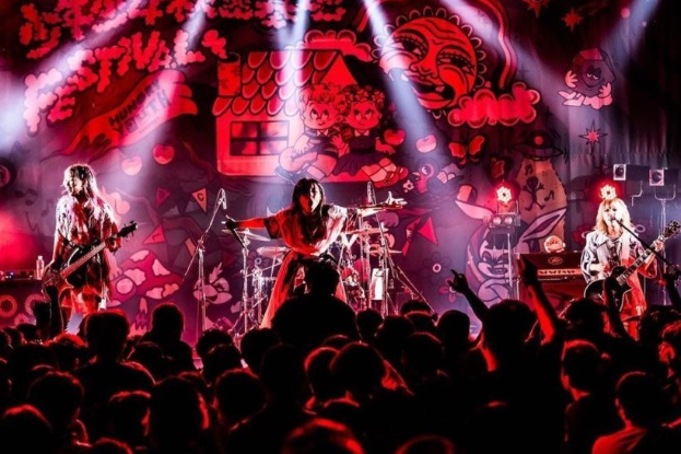 Ein Live-Auftritt der japanischen Rockband Korn auf der Bühne mit Musikern, die Gitarren spielen und singen, begleitet von Mikrofonen und Ständern, unter Bühnenlicht mit einem Gemälde im Hintergrund und einer sichtbaren Menge unten.