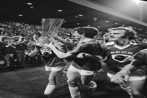 Eine Gruppe von Männern in Schwarz-Weiß-Kleidung hält einen Pokal auf einem Fußballfeld hoch und feiert ihren FA-Cup-Finalsieg, während eine Menge hinter einem Zaun und von Deckenlampen beleuchtet zusieht.