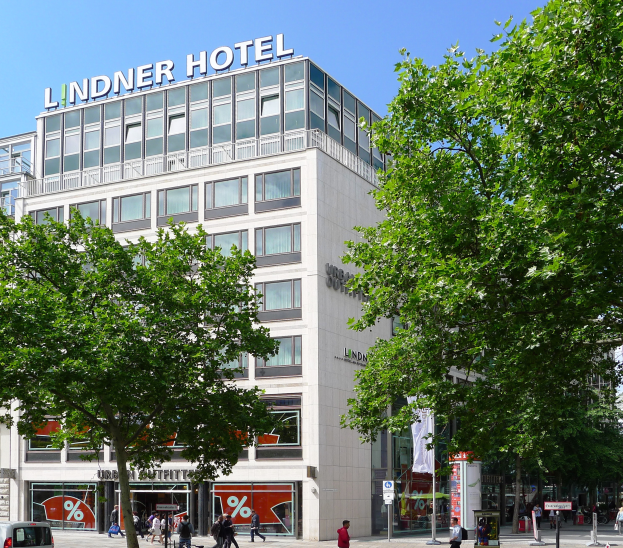 Das Lindner Hotel in Berlin, Deutschland, ein großes Gebäude mit Glasfenstern und Text an der Spitze, umgeben von Bäumen und Fahrzeugen auf der Straße, mit vielen Menschen, die herumlaufen, einige tragen Taschen, und der Himmel ist im Hintergrund sichtbar.