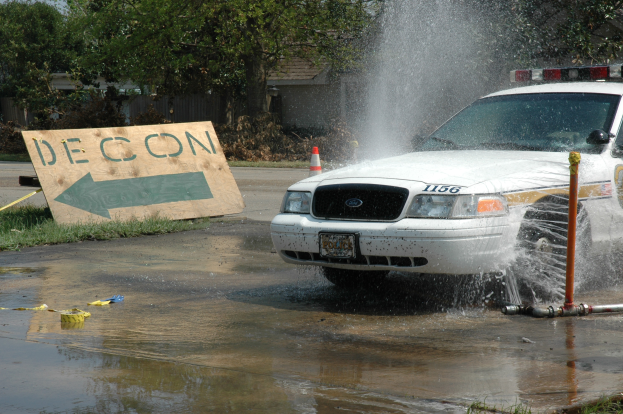 Ein Polizeiauto fährt durch eine Pfütze, mit einem Schild, einem Verkehrskegel und anderen Gegenständen auf dem Boden, vor einem Hintergrund aus Bäumen, Gebäuden und einem klaren blauen Himmel.