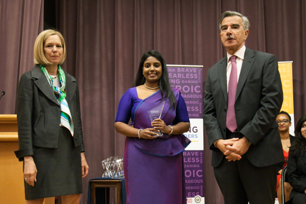 Zwei Frauen und ein Mann stehen auf einer Bühne mit einem braunen Vorhang dahinter; die Frau in der Mitte hält einen Preis, trägt ein lila Kleid und der Mann rechts trägt eine schwarze Jacke, weißes Hemd und eine pinkfarbene Krawatte; ein Podium mit einem Mikrofon ist links und mehrere Preise sind auf dem Tisch vor ihnen.