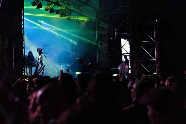Eine Menge schaut einem Live-Konzert mit Musikern auf der Bühne zu, die Instrumente spielen und unter Scheinwerfern auf der Bühne zu sehen sind, mit einem Banner, Metallrahmen und Mikrofonen.