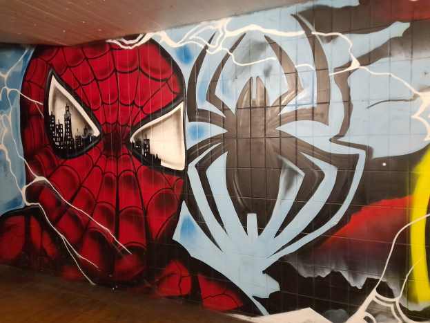 Ein buntes Wandgemälde von Spider-Man in rot und blau mit einem Webmuster und einem Spider-Logo, dargestellt auf der Seite eines Gebäudes mit einer Etage darunter und einer Decke darüber.