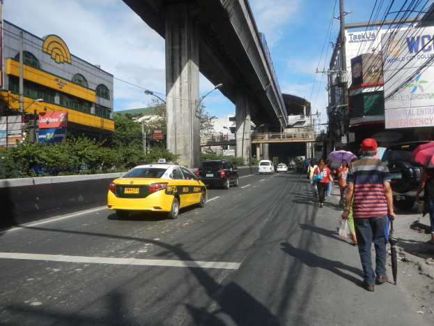 Ein gelbes Taxi fährt eine Straße mit hohen Gebäuden entlang, Fußgänger mit Regenschirmen gehen auf dem Gehweg, und in der Ferne ist eine Brücke zu sehen.