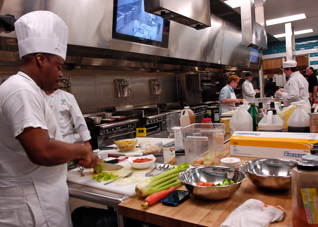 Eine Gruppe von Köchen in einer Küche, die Lebensmittel auf einem Schneidbrett zubereiten, mit einem Koch im Vordergrund in einer weißen Schürze und Mütze, der ein Messer hält und von verschiedenen Utensilien, Schüsseln, Gemüse, einem Handy, Flaschen und anderen Gegenständen umgeben ist, mit einem Fernseher und Deckenlampen im Hintergrund.