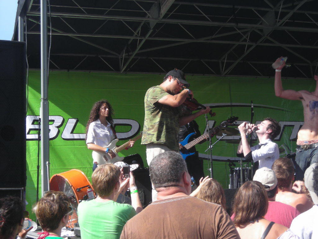 Eine Gruppe Musiker, die verschiedene Instrumente spielen, auf einer Bühne mit einem großen schwarzen Lautsprecher links, während das Publikum davor steht und die Vorstellung genießt, mit einer grünen Fahne im Hintergrund.