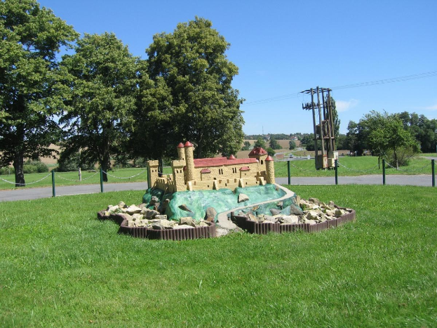 Minigolfplatz mit einem Rasenfeld, umgeben von einer Kettenzaun, Bäumen, Strommasten mit Drähten und einem bewölkten Himmel im Hintergrund.