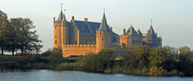 Eine Burg an einem Flussufer mit Grünfläche und einem sichtbaren Himmel im Hintergrund.