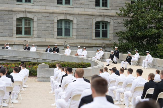 Eine Gruppe von Menschen in weißen Marineuniformen sitzt vor einem Gebäude mit Fenstern, umgeben von blühenden Pflanzen und Bäumen, während einer Abschlussfeier.