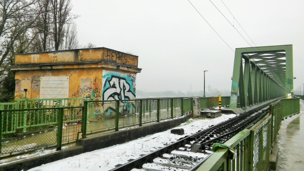Eisenbahnschiene mit Graffiti an der Seite, umgeben von Geländern, Pfählen, Lampen, Kabeln und Bäumen, mit Schnee bedeckt und sichtbarem Himmel im Hintergrund.