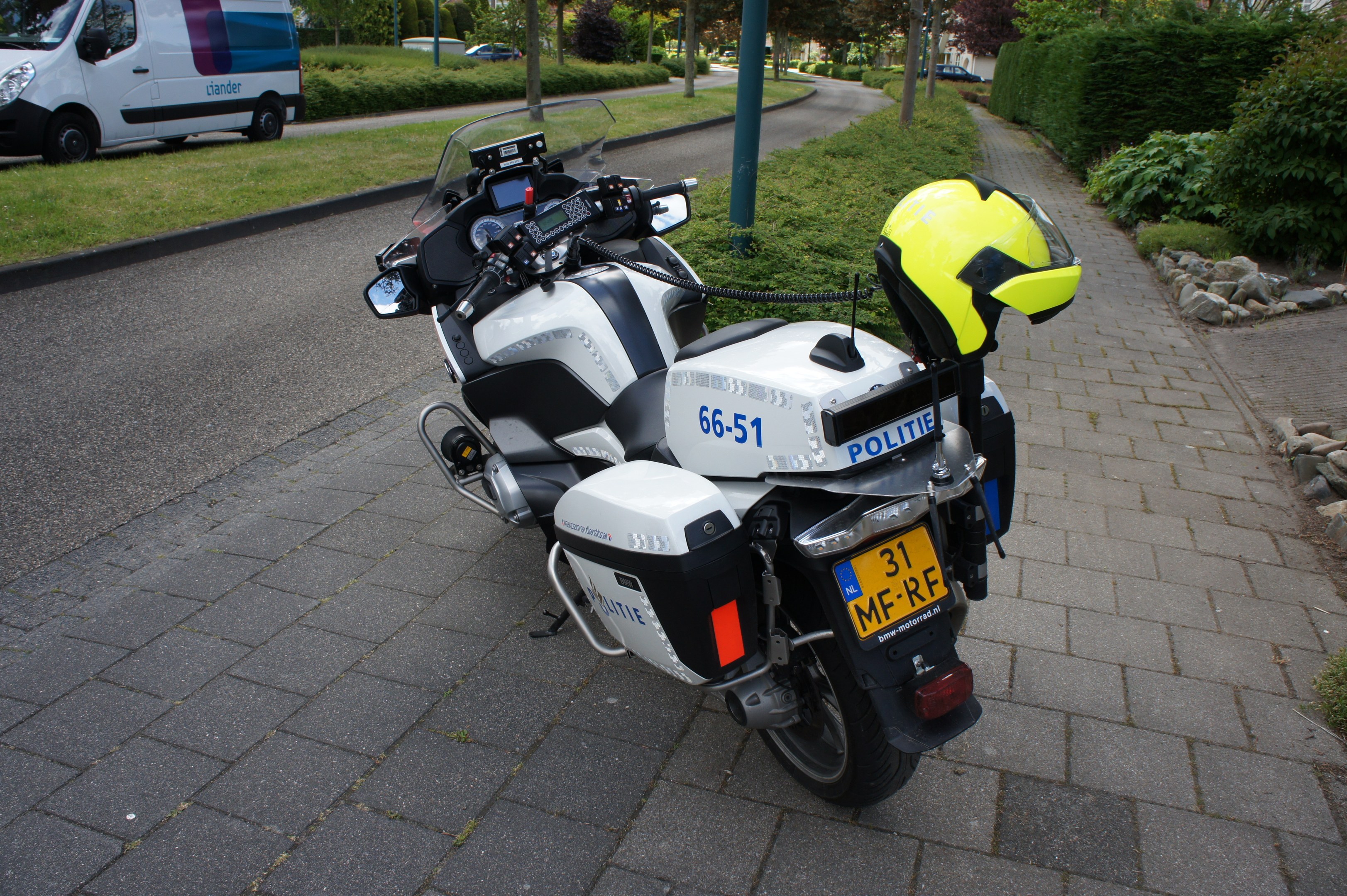 Ein polizeiliches Motorrad am Straßenrand geparkt mit einem Helm darauf, umgeben von Bäumen, Pfählen, Gebäuden, Pflanzen, Gras, Steinen und anderen Fahrzeugen im Hintergrund.
