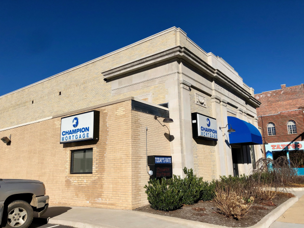 Ein zweistöckiges Gebäude mit vernagelten Fenstern, einem geparkten Auto, Topfpflanzen und Bäumen unter einem bewölkten Himmel, mit einer Tafel mit der Aufschrift "Champion Mortgage, Inc." vorne drauf.