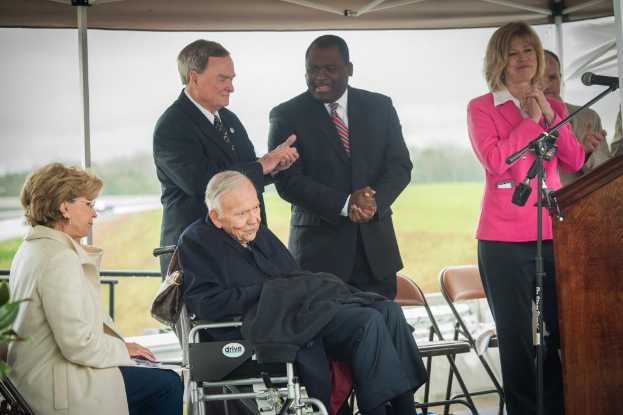 Zwei Personen sitzen unten im Bild, einige Personen stehen dahinter, Stühle, Gras und Bäume im Hintergrund, ein Podium mit Mikrofon in der rechten unteren Ecke und ein Zelt oben im Bild, sowie eine Pflanze in der linken unteren Ecke.