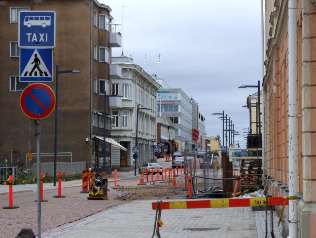 Stadtstraße mit Gebäuden, Straßenlaternen, Verkehrsschildern, Verkehrskegeln, Fahrzeugen, Absperrpoller, Bäumen, einer Baustelle mit Verkehrsschildern und einem bewölkten Himmel.