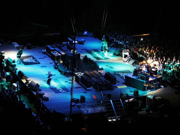 Eine Gruppe von Menschen, die auf einer Bühne mit Lautsprechern, Scheinwerfern und Mikrophonen performs, umgeben von einer großen Menge in einem dunklen Hintergrund.