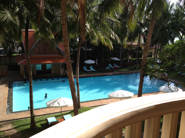 Ein großes Schwimmbad umgeben von Grün, mit Menschen, die schwimmen, Sonnenschirmen, Stühlen, Häusern im Hintergrund und einem klaren blauen Himmel, von einem Hotelbalkon aus gesehen.