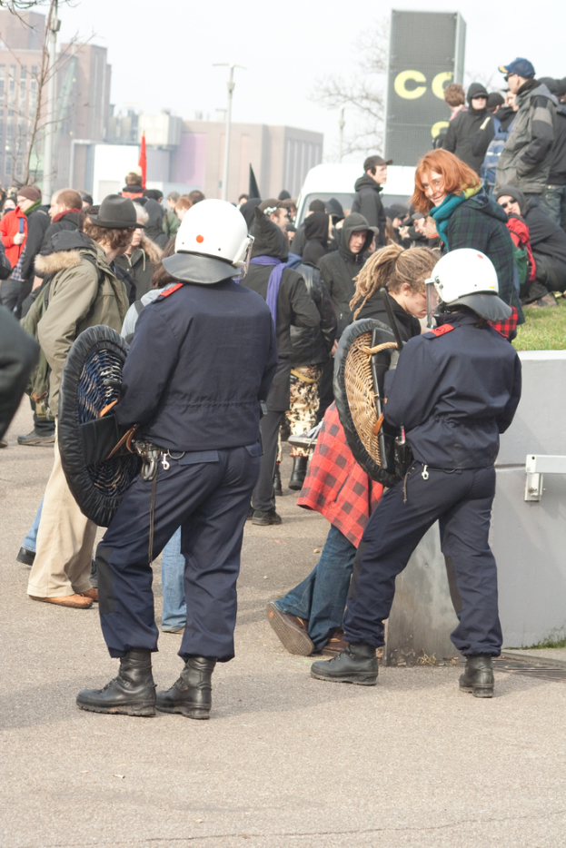 Eine Gruppe von Menschen, die auf einer Straße mit zwei Personen in der Mitte, die wie Polizeibeamte aussehen, und Gebäuden im Hintergrund gehen.