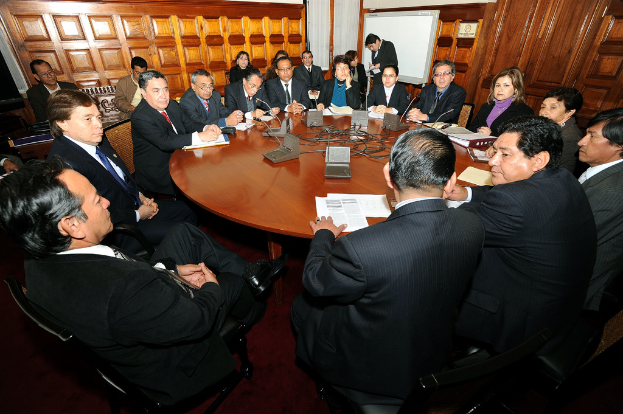 Eine Runde Tischbesprechung mit Delegierten, die über Mikrofone, Papiere, Kabel und Dateien auf dem Tisch diskutieren, mit einer hölzernen Wand im Hintergrund.