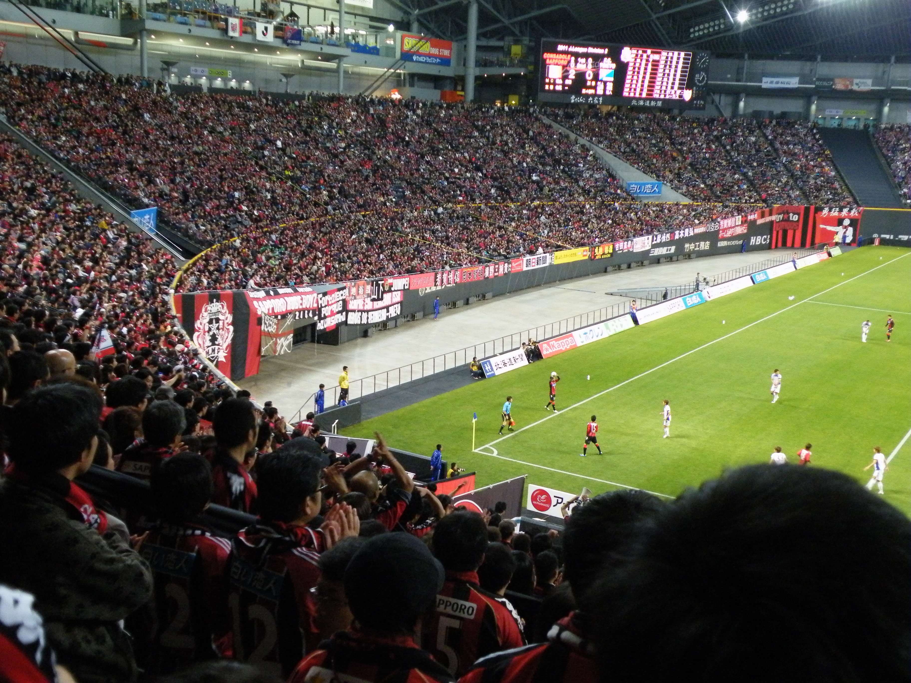 Ein Fußballspiel in einem großen Stadion mit Spielern auf dem Feld und Zuschauern in den Rängen, umgeben von Bannern, einem Zaun, Anzeigetafeln, Metallstangen und einem Dach mit Deckenbeleuchtung.