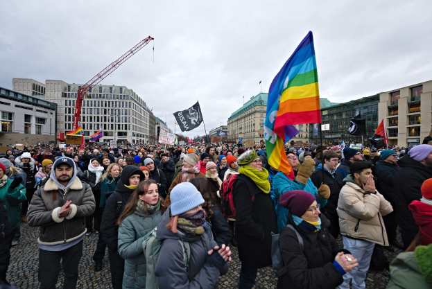 Große Gruppe von Menschen mit LGBTQ+-Rechten-Bannern und -Fahnen vor einem Gebäude mit einem Kran und einem bewölkten Himmel.