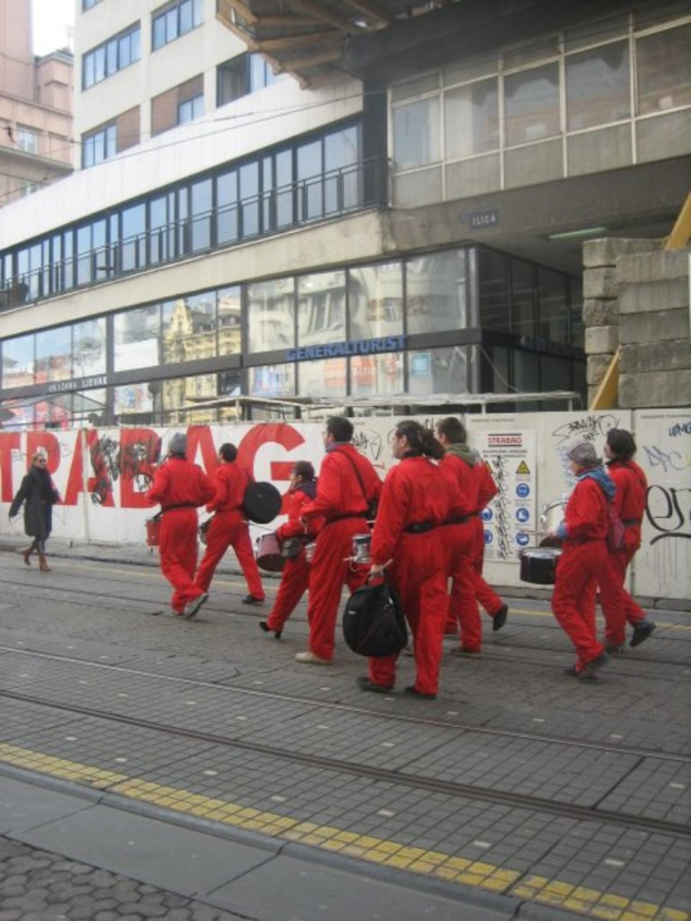 Menschen, die auf einem Boden gehen und Musikinstrumente spielen, mit einer Umzäunungswand dahinter, die Plakate und Text trägt, und weiter hinten Gebäude mit Glaswänden und Fenstern, von denen einige Namen tragen.