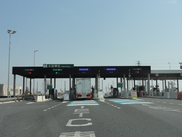 Eine Mautstation am Straßenrand mit einem vor ihr geparkten Lkw, weiße und blaue Straßenmarkierungen, Laternenpfähle, Schilder und andere Gegenstände auf beiden Seiten, mit Gebäuden, Bäumen und einem klaren blauen Himmel im Hintergrund.