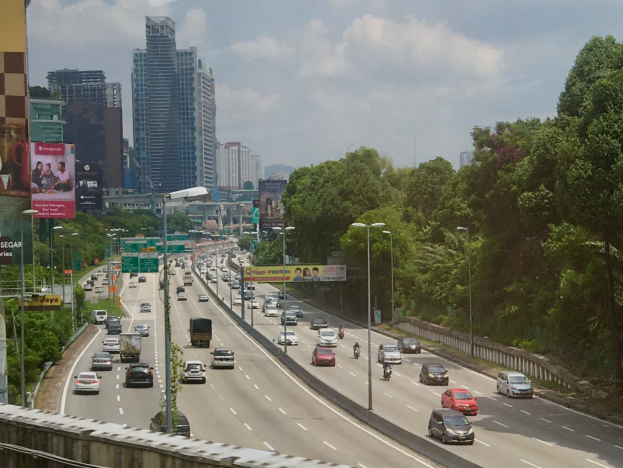 Eine stark befahrene Autobahn mit zahlreichen Fahrzeugen, gesäumt von hohen Gebäuden, Straßenlaternen, Werbetafeln und Bäumen unter einem bewölkten Himmel.
