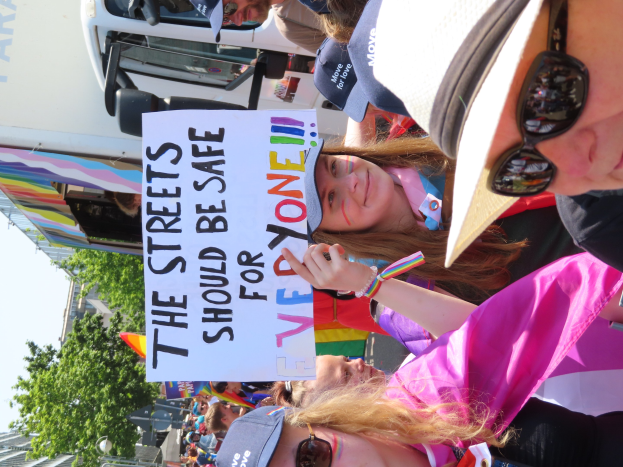 Eine Gruppe von Menschen steht vor einem Lastwagen und hält ein Schild mit der Aufschrift "Die Straßen sollten sicher sein", einige tragen Mützen und Schutzbrillen und andere halten Schilder, vor einem Hintergrund aus Bäumen, Gebäuden und einem klaren blauen Himmel.