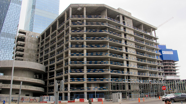 Großes Bauwerk im Bau in einer Stadt umgeben von Wolkenkratzern, Fahrzeugen, Fußgängern, Verkehrsinfrastruktur und Kränen vor einem klaren blauen Himmel.