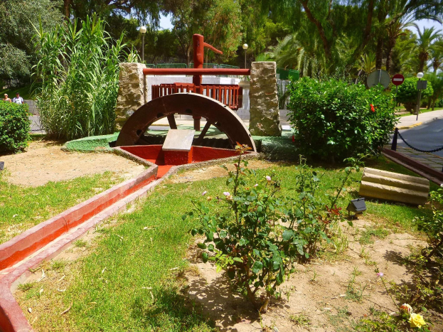 Minigolfanlage in einem Park mit Grünfläche, Straße, Fußgängern, Schildern und einem blauen Himmel.
