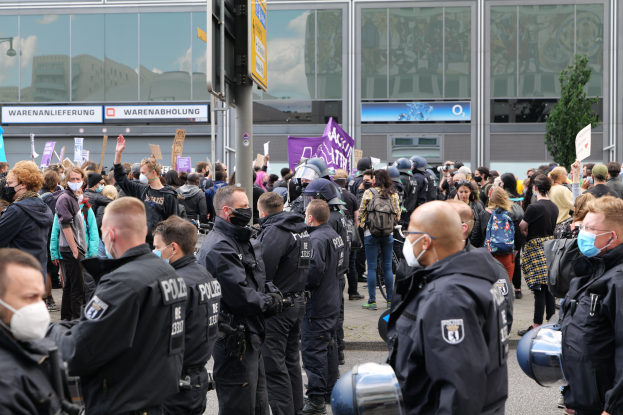 Eine große Gruppe von Menschen steht vor einem Gebäude, einige halten Schilder und tragen Helme, mit einem Mast mit einer Schautafel im Vordergrund und einem Baum im Hintergrund, scheinbar protestierend.