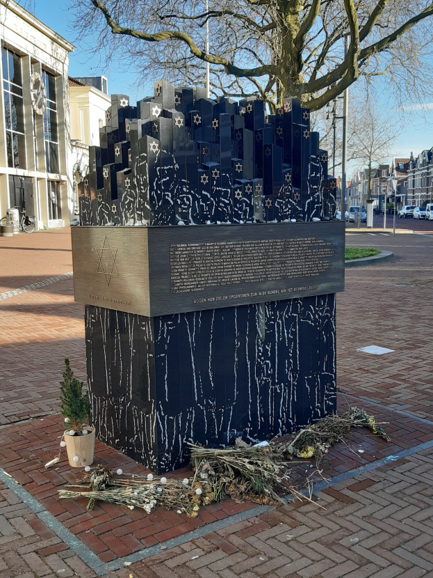 Denkmal für Holocaust-Opfer in einem Stadtplatz mit einem Baum davor, einem Topf mit einer Pflanze, verstreuten trockenen Blättern und umgebenden Gebäuden, Fahrzeugen, Masten und Bäumen.
