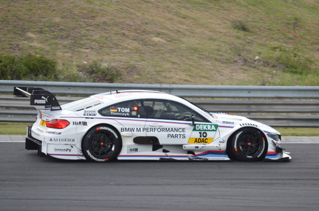 Ein BMW M4 DTM-Rennwagen fährt auf einer Rennstrecke, umgeben von saftigem Grün, mit einer Begrenzung im Hintergrund.