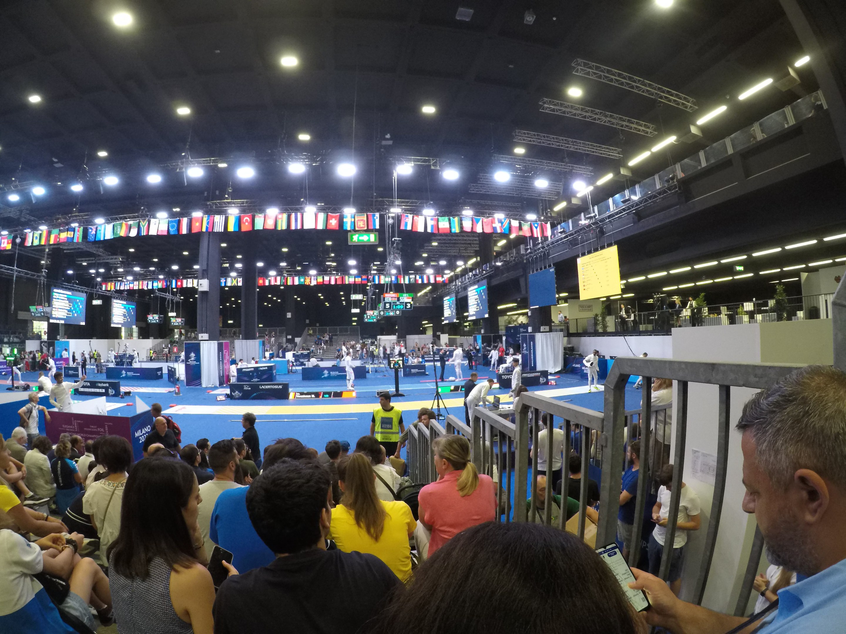 Eine große Menge Menschen sitzt und steht in einer Indoor-Sportarena mit Bildschirmen, Flaggen und Deckenbeleuchtung im Hintergrund.