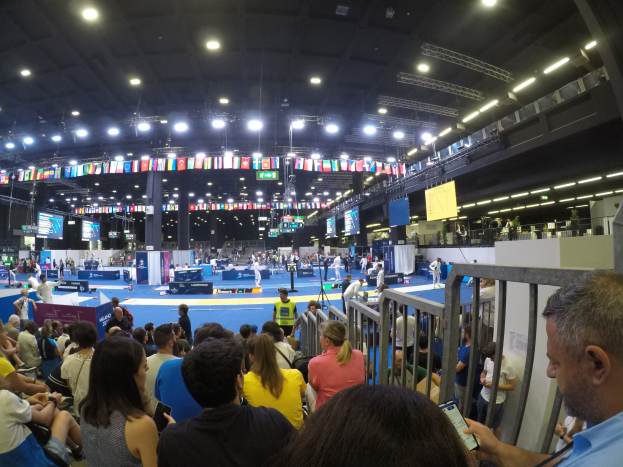 Eine große Menge Menschen sitzt und steht in einer Indoor-Sportarena mit Bildschirmen, Flaggen und Deckenbeleuchtung im Hintergrund.