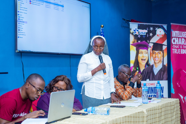 Eine Frau mit Mikrofon spricht zu einer Gruppe, die um einen Tisch mit Laptops, Papieren und Flaschen sitzt, mit einem Banner und Flaggen im Hintergrund.