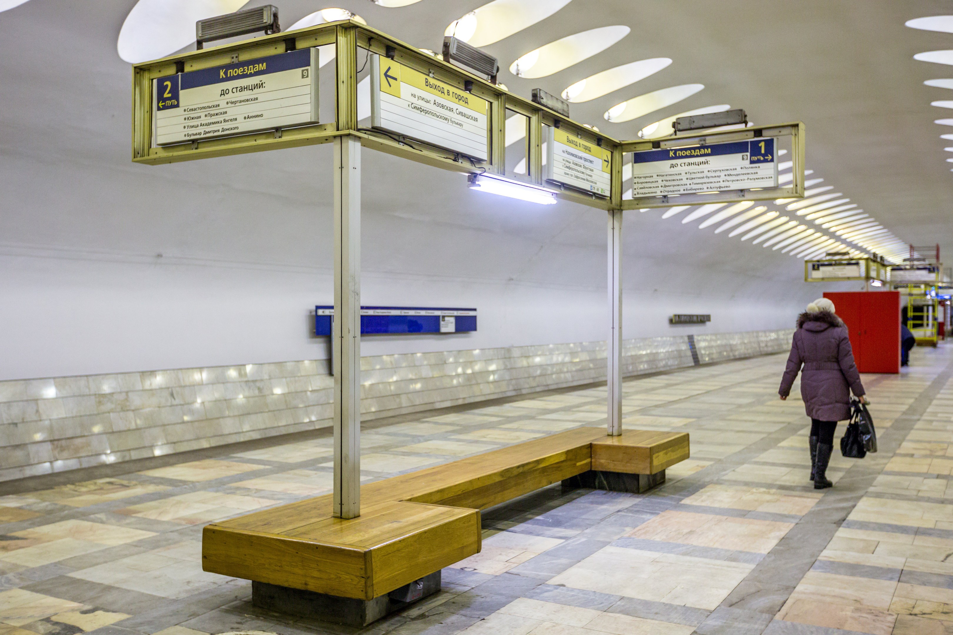 Eine Frau in einem Mantel mit einer Tasche geht durch eine belebte Moskauer U-Bahn-Station mit Bänken, Informationsanzeigen und Beleuchtung.