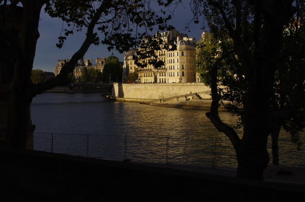 Ein Zaun, Wasser, Bäume, Gebäude, Laternenpfähle, Fahrzeuge auf einer Straße und der Himmel, möglicherweise in der Nähe eines Sees.