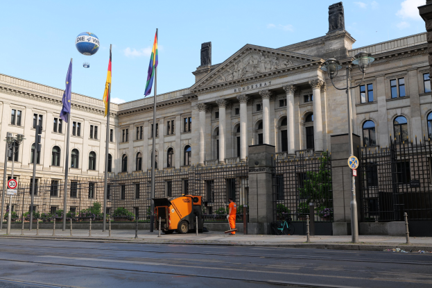 Großes modernes Gebäude mit zahlreichen Fenstern, identifiziert als der Bundestag in Berlin, Deutschland, umgeben von Straßeninfrastruktur und Fußgängern, mit Flaggen und Bäumen im Vordergrund und einem bewölkten Himmel im Hintergrund.